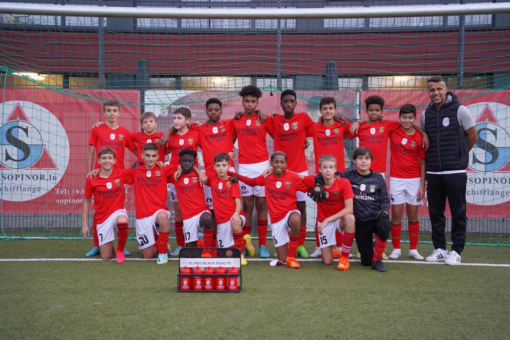 Eine Gruppe junger Männer in roten T-Shirts und schwarzen Shorts steht auf einem Fußballfeld vor einer Kiste roter Gegenstände, im Hintergrund Schilder mit der Aufschrift "FC Red Black Eglaute - Champions League"