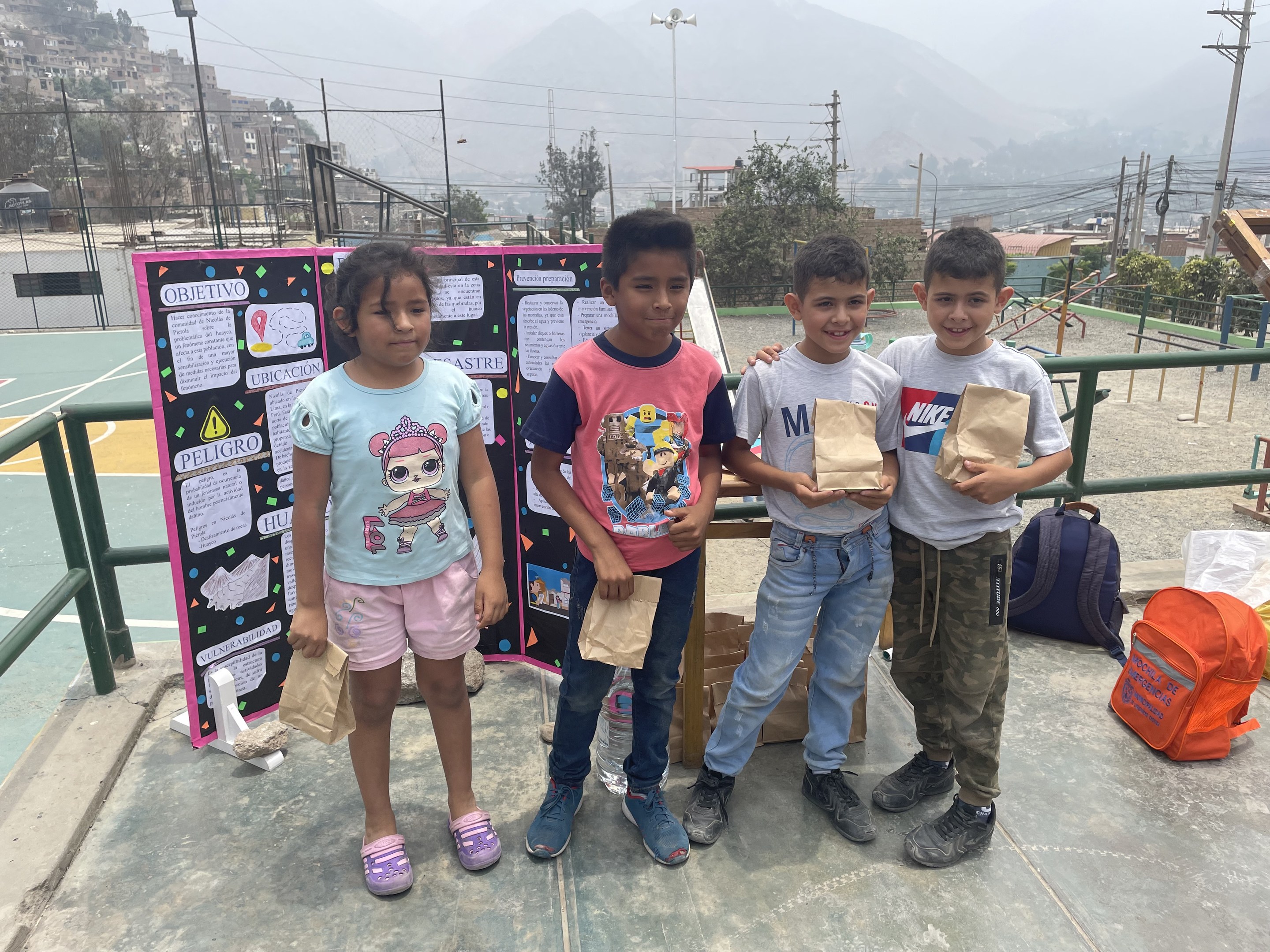 Gruppe von Kindern auf einem Basketballfeld, die Papierbeutel in den Händen halten, mit Beuteln und Gegenständen rechts daneben und einer Tafel mit Text links daneben, vor einer Außenkulisse mit Zaun, Pfosten, Laternen, Strommasten, Bäumen, Gebäuden, Hügeln und Himmel.