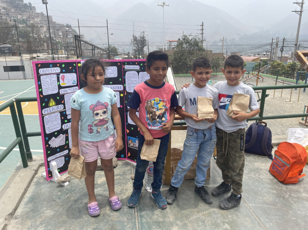 Gruppe von Kindern auf einem Basketballfeld, die Papierbeutel in den Händen halten, mit Beuteln und Gegenständen rechts daneben und einer Tafel mit Text links daneben, vor einer Außenkulisse mit Zaun, Pfosten, Laternen, Strommasten, Bäumen, Gebäuden, Hügeln und Himmel.