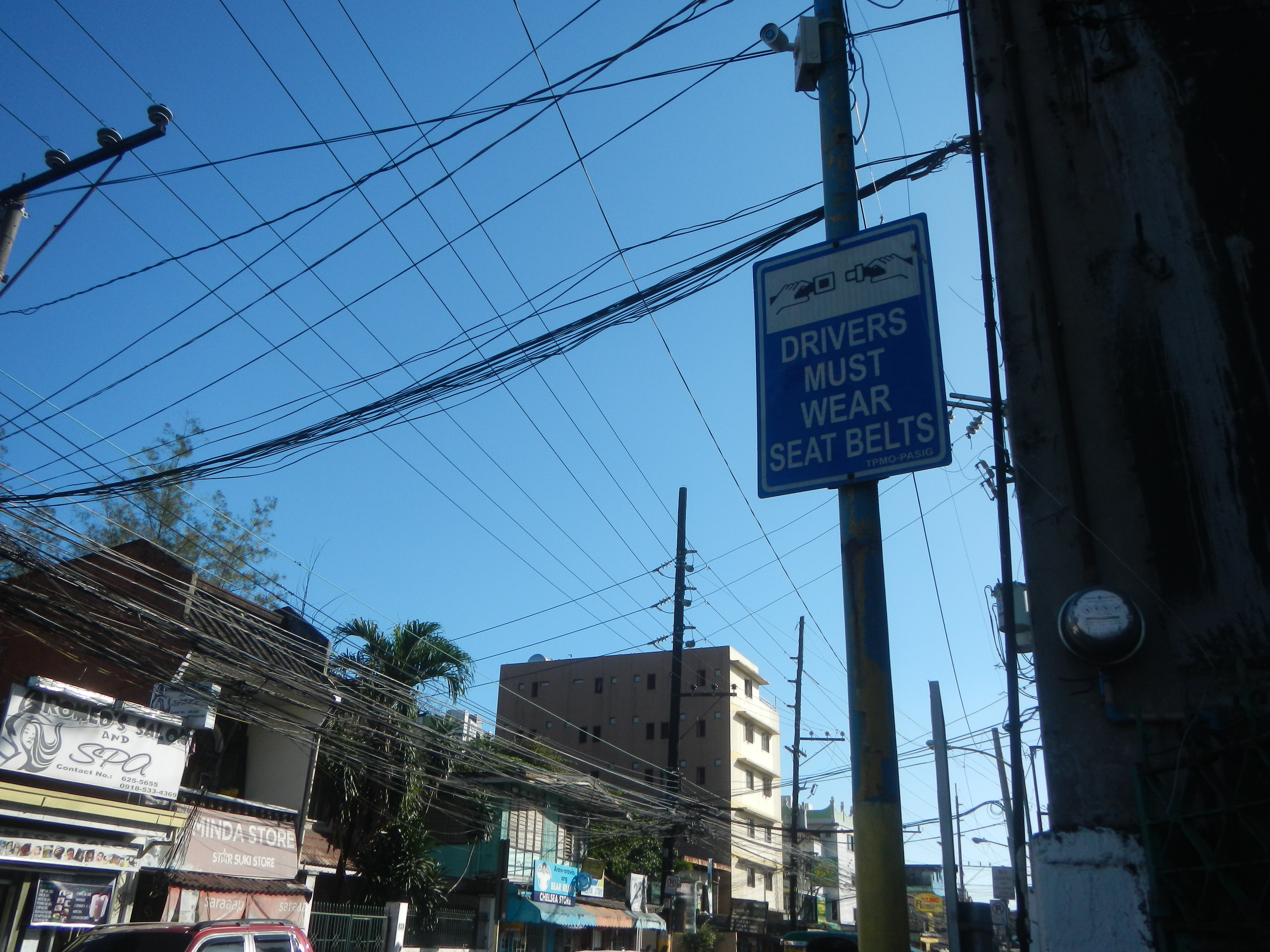 Eine Stadtstraße mit Autos, Strommasten mit Drähten, Gebäuden, Bäumen und Namensschildern, mit einem Schild an einem Mast, auf dem "Fahrer müssen Sicherheitsgurte tragen" steht und ein sichtbarer Himmel im Hintergrund.