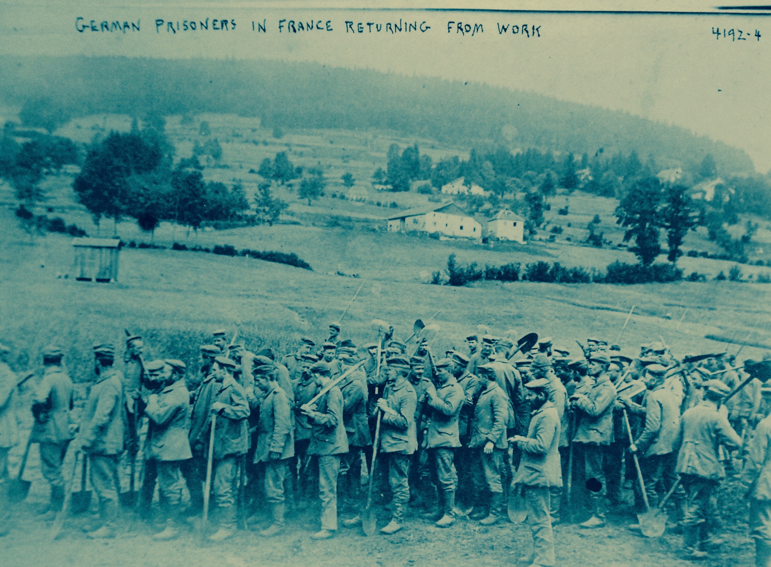 Eine Gruppe deutscher Gefangener in Frankreich, die Gewehre in der Hand halten, steht auf dem Boden mit Bäumen, Häusern und einem klaren blauen Himmel im Hintergrund.