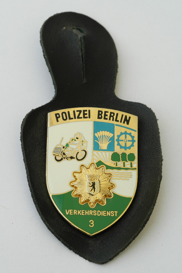 Ein schildförmiges Polizewemzeichen mit "Polizei Berlin" in schwarzer fettgeschriebener Schrift auf einem blauen Hintergrund mit einem weißen Stern und Rahmen, das auf einer Oberfläche ruht.