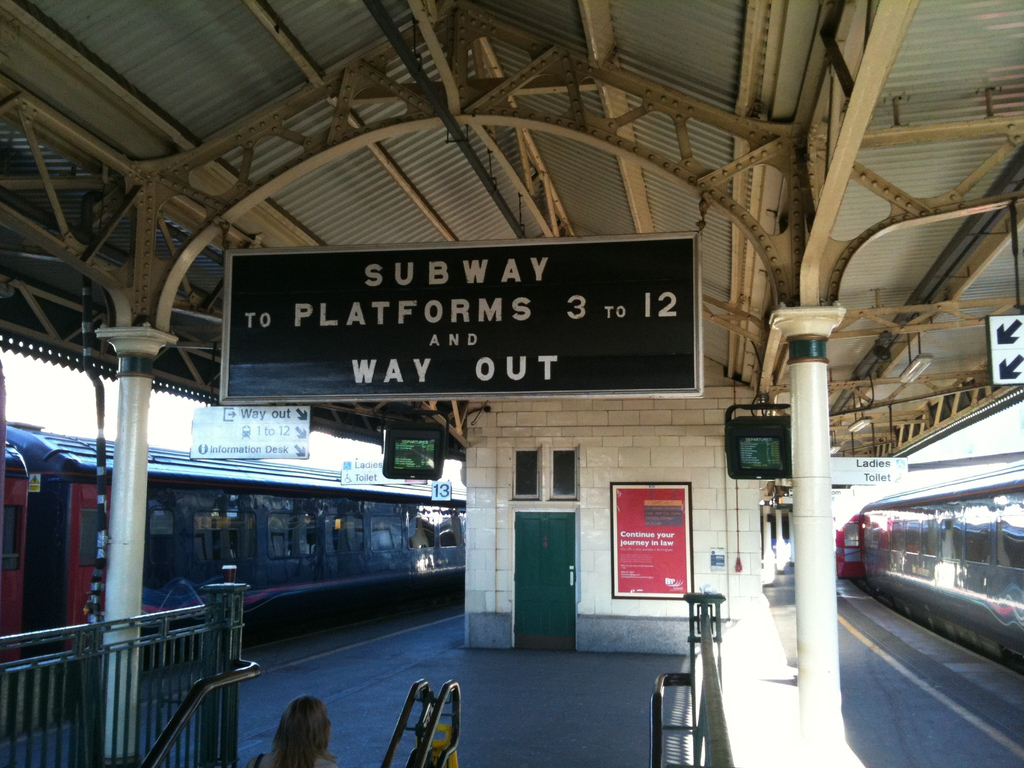 Zwei Züge stehen neben einem Bahnsteig mit zwei Bildschirmen und mehreren Schildern, die von der Decke hängen, eine Person ist in der Nähe der Treppe und eine Tür ist an der Wand sichtbar.