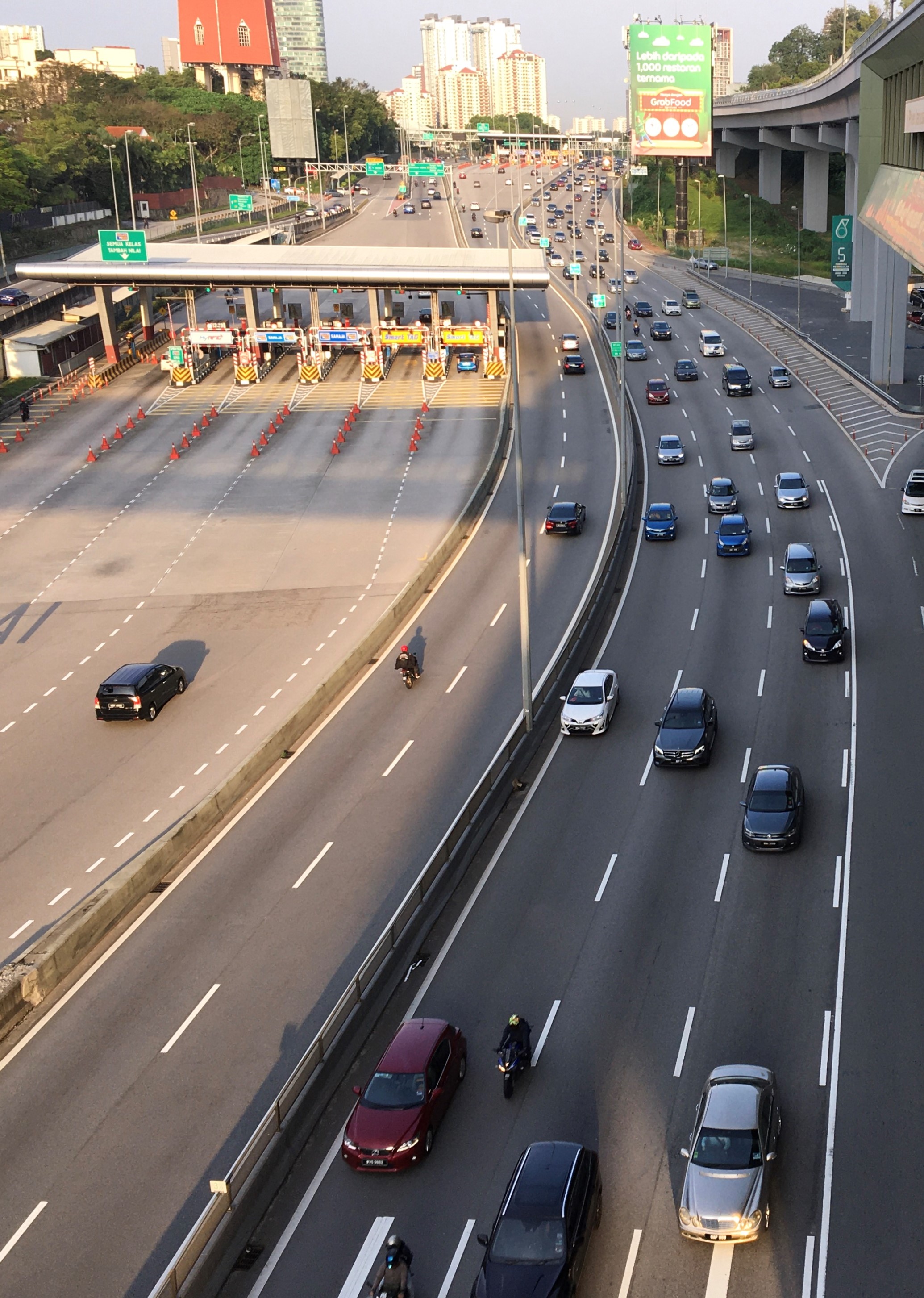 Eine belebte Autobahn mit zahlreichen Autos, Motorrädern, Verkehrskegeln, Verkehrszeichen, Laternenmasten, einer Brücke, Bäumen, Gebäuden und einem klaren blauen Himmel.