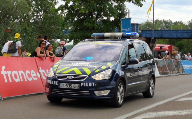Polizeiauto fährt auf einer Straße neben einer Menschenmenge, mit einer Fahne auf der linken Seite, Geländern mit Fahnen dahinter, Bäumen, einer Brücke, einer Fahne auf einem Mast und einem bewölkten Himmel im Hintergrund.