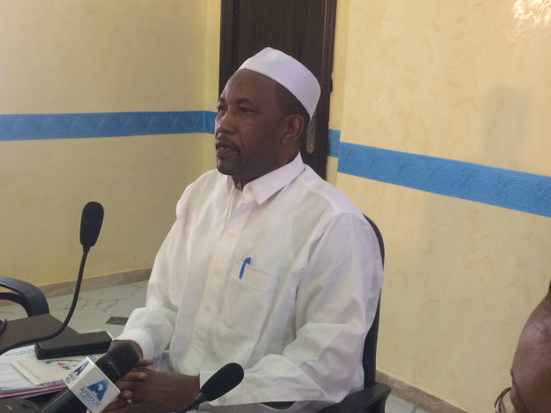 Ein Mann in weißem Anzug und Hut sitzt an einem Tisch mit Mikrofonen, spricht, während Papiere und Gegenstände auf dem Tisch liegen, mit einem anderen Mann mit Brille rechts von ihm in einem Raum mit Wand und Tür im Hintergrund.