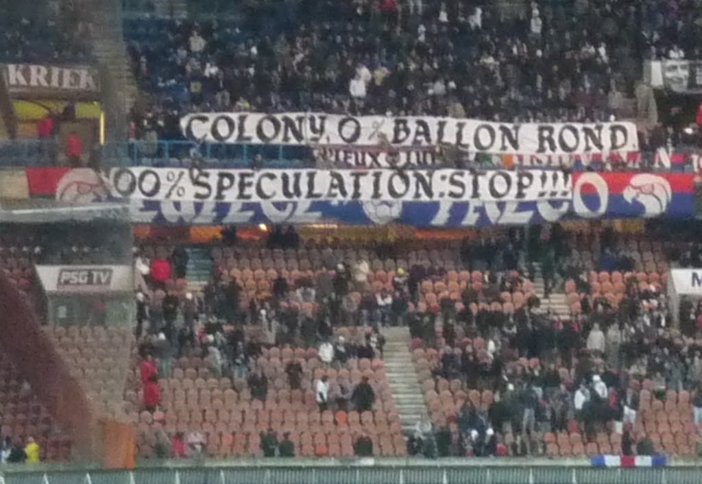 Ein Stadion voller Menschen, Fahnen und Stühle, mit einem Zaun unten und einer Treppe links.