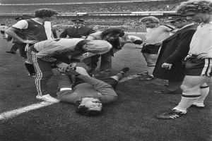 Schwarzes und weißes Bild eines Fußballspielers, der auf dem Boden liegt und von einer Gruppe von Männern umgeben ist, mit einer Menge im Stadionhintergrund; der Spieler scheint verletzt zu sein.