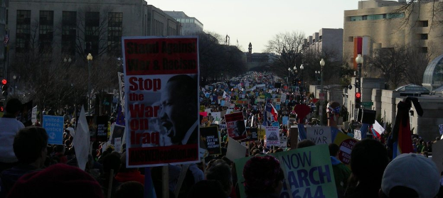Menschen auf einer Straße mit Plakaten, im Hintergrund Gebäude, Bäume, Laternenpfähle und der Himmel.