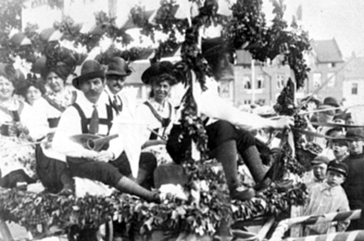 Schwarzes und weißes Bild von Menschen in traditioneller bayrischer Tracht auf einem geschmückten Festwagen in einem Umzug, einige spielen Instrumente, mit Gebäuden und einem klaren Himmel im Hintergrund.