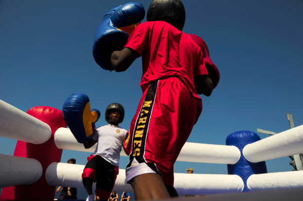 Zwei Boxer in einem Boxring, einer in rotem Shirt und blauen Shorts, kämpfen mit Boxhandschuhen, umgeben von ein paar Menschen und unter einem klaren blauen Himmel.