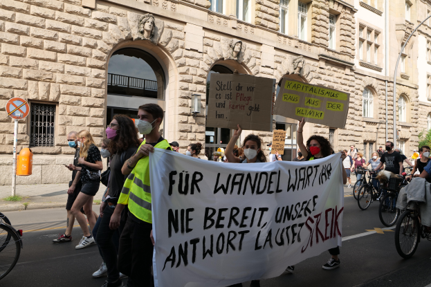 Eine Gruppe von Menschen marschiert auf der Straße in Berlin, hält Schilder und Banner hoch und fährt mit Fahrrädern, vor einem Gebäude mit Fenstern, Bögen, Säulen, Skulpturen und Bäumen.