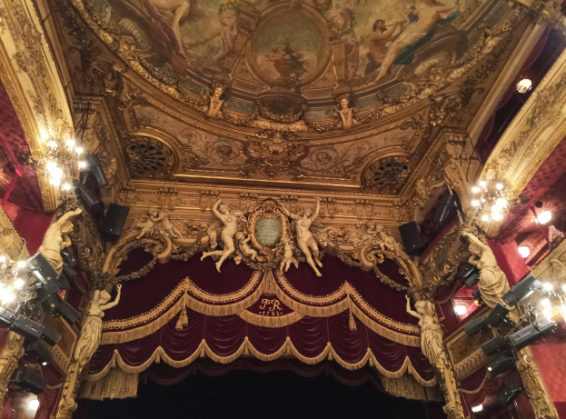 Theaterinnenraum mit roten Vorhängen, Kronleuchtern, Skulpturen, Lichtern und einer bemalten Decke, mit einer Bühne und einer kleinen Gruppe von Menschen davor.