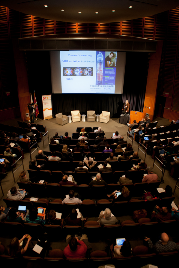 Ein mit sitzenden Menschen gefüllter Hörsaal, der Gegenstände hält, von vielen Lichtern beleuchtet, mit Fahnen links, einem Podium rechts und einer Werbetafel links.