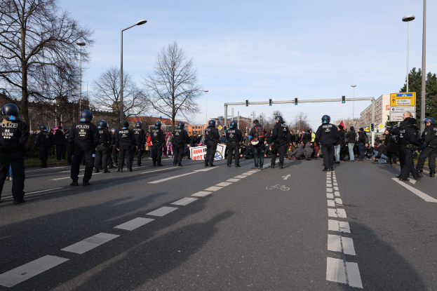 Gruppe von Polizisten in schwarzen Uniformen und Helmen auf der Seite einer Straße mit Laternenpfählen, Verkehrsampeln, Bäumen, Gebäuden und einem klaren blauen Himmel im Hintergrund.