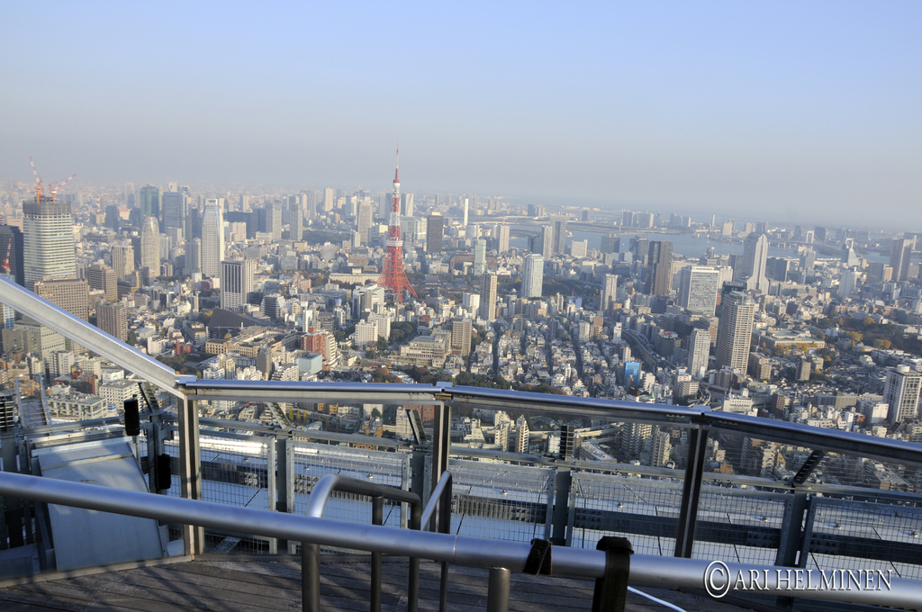 Luftaufnahme einer Stadt mit Gebäuden, einem Fluss, einem Metallstabzaun im Vordergrund und Text unten.