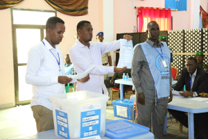 Eine Gruppe von Männern steht um einen Tisch mit Kisten, Papieren und anderen Gegenständen, die Stimmzettel ausfüllen, im Hintergrund sind Menschen auf Stühlen und einige stehen auf dem Boden, Tische, Flaschen, Vorhänge, eine Tür, eine Wand, eine Klimaanlage und eine Flagge zu sehen.