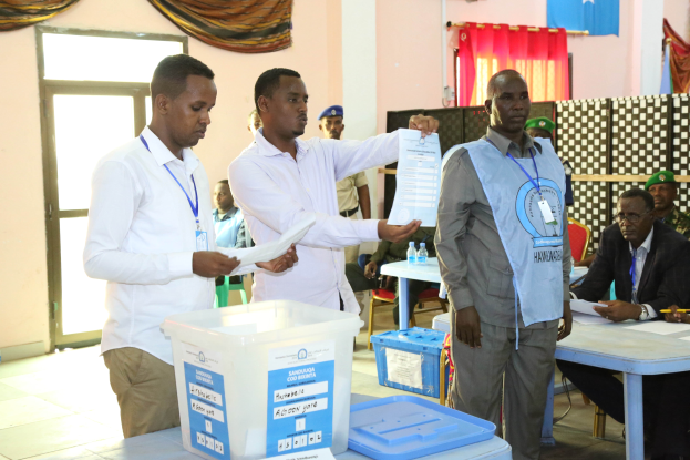 Eine Gruppe von Männern steht um einen Tisch mit Kisten, Papieren und anderen Gegenständen, die Stimmzettel ausfüllen, im Hintergrund sind Menschen auf Stühlen und einige stehen auf dem Boden, Tische, Flaschen, Vorhänge, eine Tür, eine Wand, eine Klimaanlage und eine Flagge zu sehen.