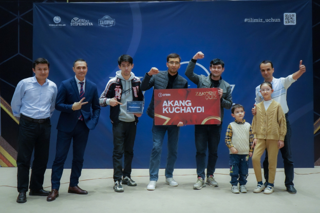 Gruppe von Menschen auf einer Bühne, die eine Tafel halten, auf der "Akang Kuchaydi - Zakovat Quiz" steht, mit einem Banner und Lichtern im Hintergrund.