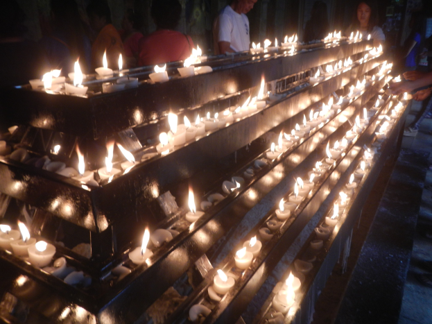 Eine Gruppe von Menschen steht um eine lange Reihe von Kerzen in einer Kirche, beleuchtet von flackerndem Kerzenlicht vor einer Wand.