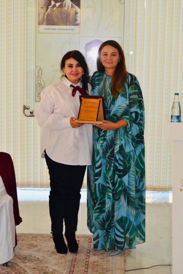 Zwei Frauen bei einer Preisverleihung, eine in einem weißen Hemd und schwarzen Hosen mit einer Auszeichnung, die andere neben einem Rednerpult mit einer Wasserflasche, mit Vorhängen und einem Fotorahmen im Hintergrund.
