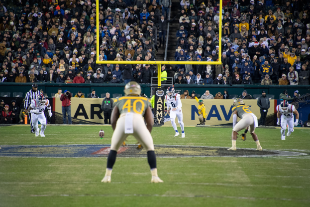 Fußballspieler in Sportbekleidung und Helmen konkurrieren auf einem Feld mit Zuschauern im Hintergrund, Stadionstrukturen und sichtbarer Beschilderung.