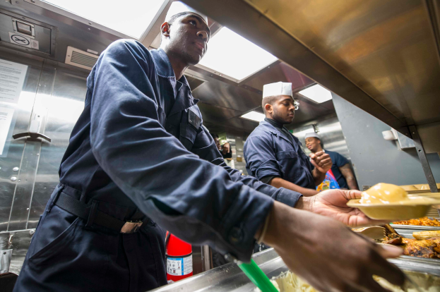 Eine Gruppe von Männern steht in einer Küche, einer hölt eine Speisetafel, mit Tabletts voller Speisen auf dem Tisch, einem Feuerlöscher und Papieren an der Wand im Hintergrund.