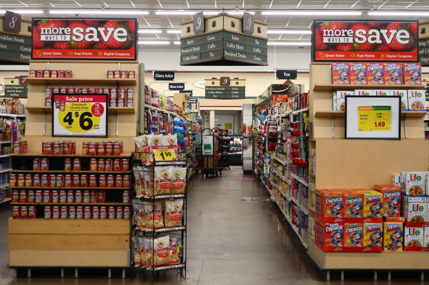Ein Convenience-Store-Innenraum mit Regalen und Boden voller verschiedener Lebensmittel, Deckenbeleuchtung und einer Person im Hintergrund.