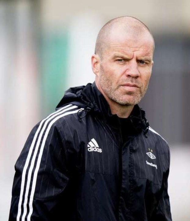 Ein kahlköpfiger Mann in einer schwarzen Jacke steht auf einem Fußballfeld und trägt eine marineblaue Jacke mit weißen Streifen und einem weißen Kragen. Seine Hände sind verschränkt und er hat einen ernsten Gesichtsausdruck.