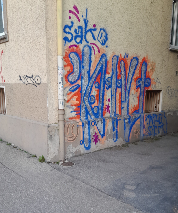Ein Gebäude mit Fenstern, einem Rohr und Graffiti an der Seite, mit einer Straße am unteren Bildrand.
