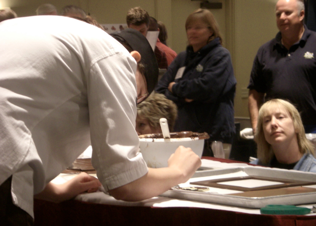 Eine Gruppe von Menschen vor einem Tisch mit einem Löffel, einer Schüssel und einem Tablett darauf, mit einer Bibel und einer Wand im Hintergrund.