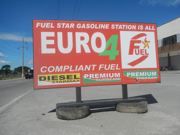 Eine Tankstelle mit einem "Euro4 Fuel"-Schild im Vordergrund, Fahrzeuge auf der Straße, Strommasten mit Drähten, Bäume, ein Gebäude auf der rechten Seite und sichtbarer Himmel im Hintergrund.
