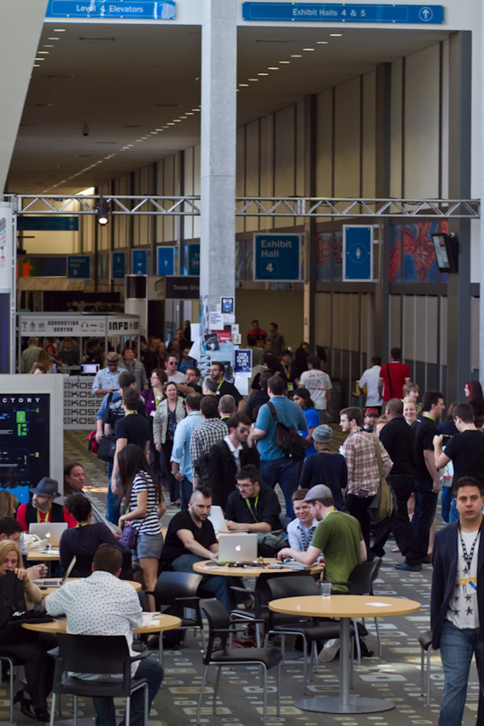 Ein belebter Flughafenvorraum mit wartenden Menschen, einige stehen und andere arbeiten, Richtungsschilder an der rechten Wand und darüber, sowie von der Decke hängende Lampen.