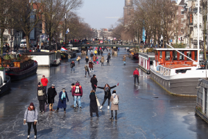 Eine Gruppe von Menschen, die auf einem zugefrorenen Kanal in Amsterdam Eislaufen, umgeben von Booten, Bäumen, Gebäuden mit Fenstern, Laternen, Flaggen und einer Brücke unter einem klaren Himmel.