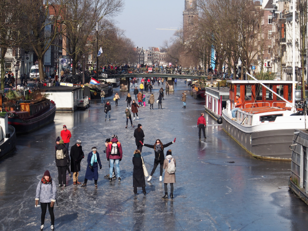 Eine Gruppe von Menschen, die auf einem zugefrorenen Kanal in Amsterdam Eislaufen, umgeben von Booten, Bäumen, Gebäuden mit Fenstern, Laternen, Flaggen und einer Brücke unter einem klaren Himmel.