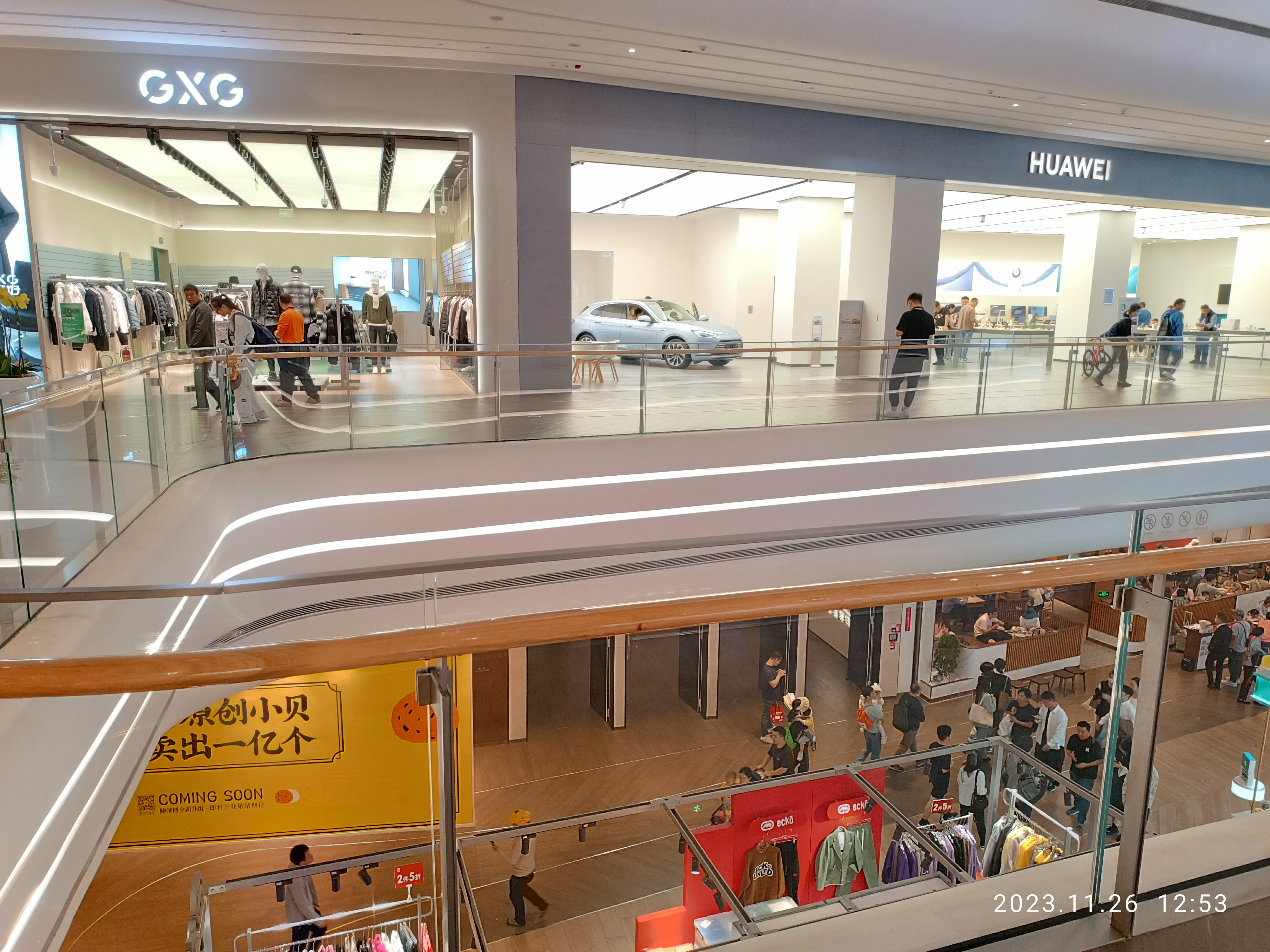 Ein Einkaufszentrumsinneres mit Menschen, die gehen, Glasgeländern, Bekleidungsauslagen, Informationsschildern, Säulen, Deckenlampen und einem geparkten Auto im Hintergrund, mit einem GXG- und Huawei-Partnerschafts-Wasserzeichen.