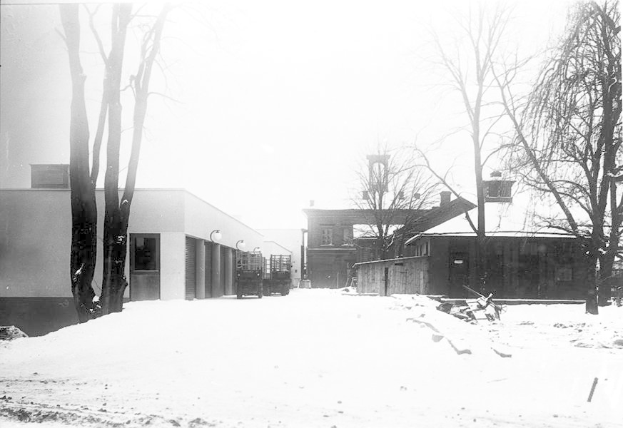 Ein Schwarz-Weiß-Foto einer verschneiten Straße mit einem Schulgebäude im Hintergrund, umgeben von Bäumen unter einem klaren Himmel.