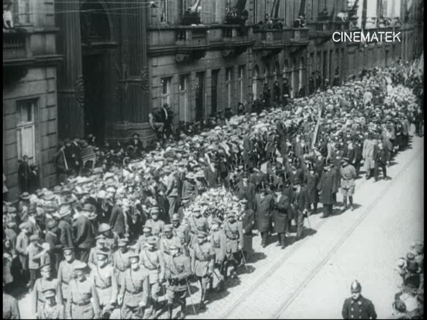 Schwarzes und weißes Foto einer großen Menge, die marschiert, einige halten Gewehre, vor einem Gebäude mit einem Wasserzeichen in der oberen rechten Ecke.