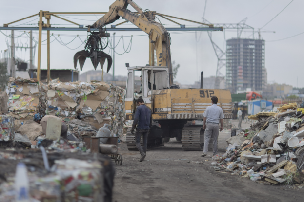 Eine Gruppe von Männern, die auf einem Schotterweg neben einem Müllhaufen gehen, mit einem Bagger im Hintergrund, Gebäuden, Bäumen, Polen und Drähten, die in der Gegend verstreut sind.