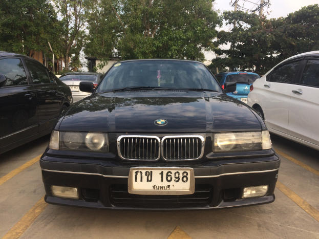 Ein schwarzer BMW E36 318i auf einem Parkplatz mit anderen Autos, sichtbares Kennzeichen, Bäume, Pfosten, Gebäude und einen klaren blauen Himmel im Hintergrund.