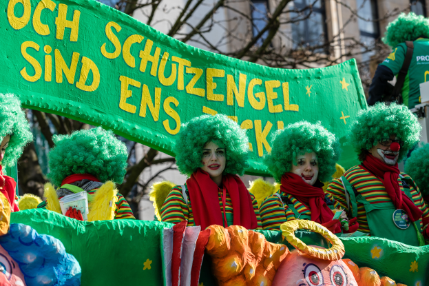 Eine Gruppe von Clowns in bunten Kostümen marschiert durch eine Straße, hält ein Schild mit Text hoch, mit Bäumen und einem Gebäude im Hintergrund.