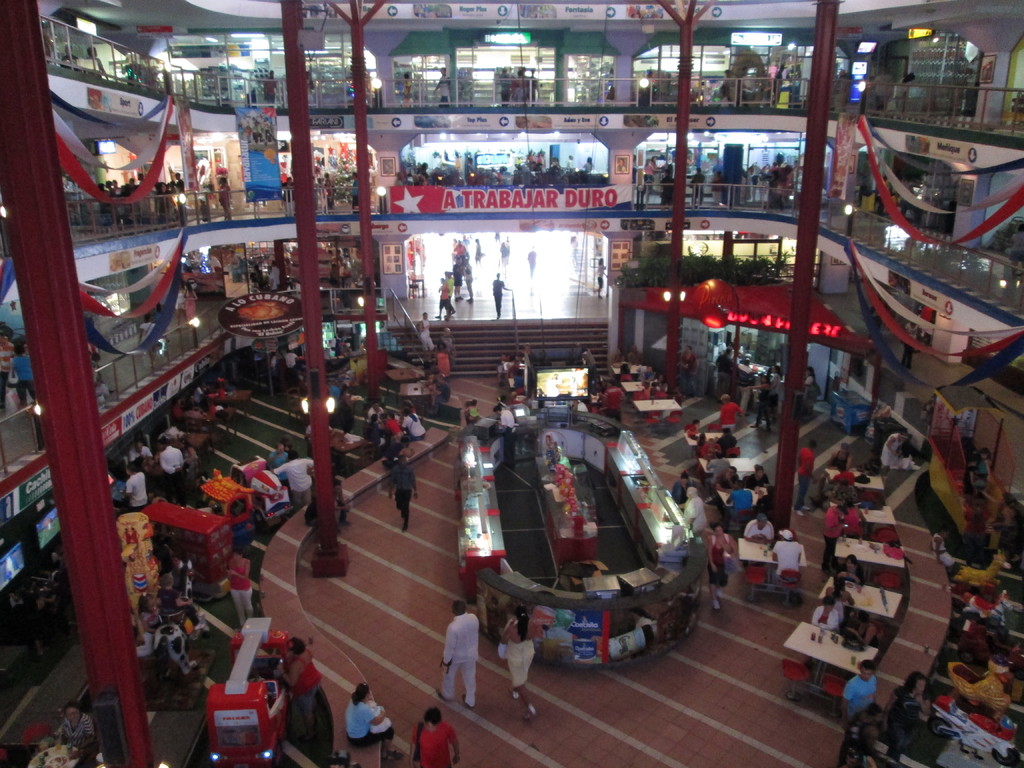 Ein belebtes Einkaufszentrum mit Säulen, Menschen, Tischen, Fliesen, Flaggen, Geschäften, einem Banner, Treppen und einem Namensschild.