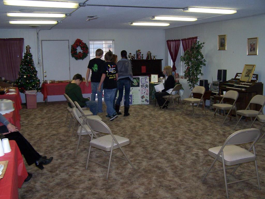 Ein Raum mit mehreren Stühlen, Tischen und Menschen, einige stehen und einige sitzen, mit Tassen und Gefäßen auf den Tischen, einem Weihnachtsbaum, einem Piano auf der rechten Seite, Foto Rahmen, Fenstern und Röhrenleuchten an der Decke und einer Wand mit einem Bouquet, Vorhang, Tür und mehr Foto Rahmen im Hintergrund.