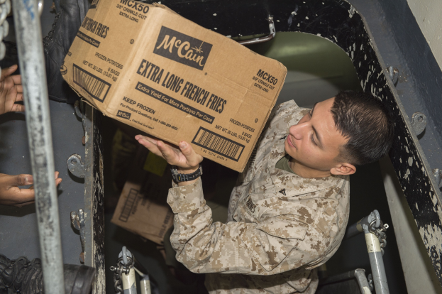Ein Mann in militärischer Uniform, der eine Kiste mit Lebensmitteln hält, mit der Hand einer anderen Person im Hintergrund, die Kiste in einen Lastwagen lädt.