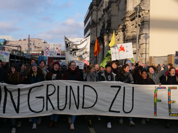 Eine Gruppe von Menschen marschiert mit einem Banner, auf dem 'In Grund zu Feu' steht, die Straße entlang und protestiert gegen das Internetverbot der deutschen Regierung. Die Straße ist von Gebäuden, Pfählen und Flaggen gesäumt.