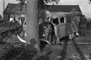 Ein Schwarz-Weiß-Bild eines verunglückten Autos am Straßenrand mit einem Baum im Vordergrund, einigen Menschen in der Nähe und Häusern, Bäumen und einem Pfahl im Hintergrund.