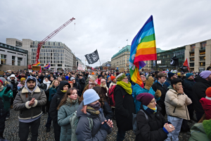 Eine große Gruppe von Menschen auf einer LGBTQ+-Rechtsdemo in Berlin, die Fahnen und Plakate schwenken, mit Gebäuden, einem Kran und einem bewölkten Himmel im Hintergrund.