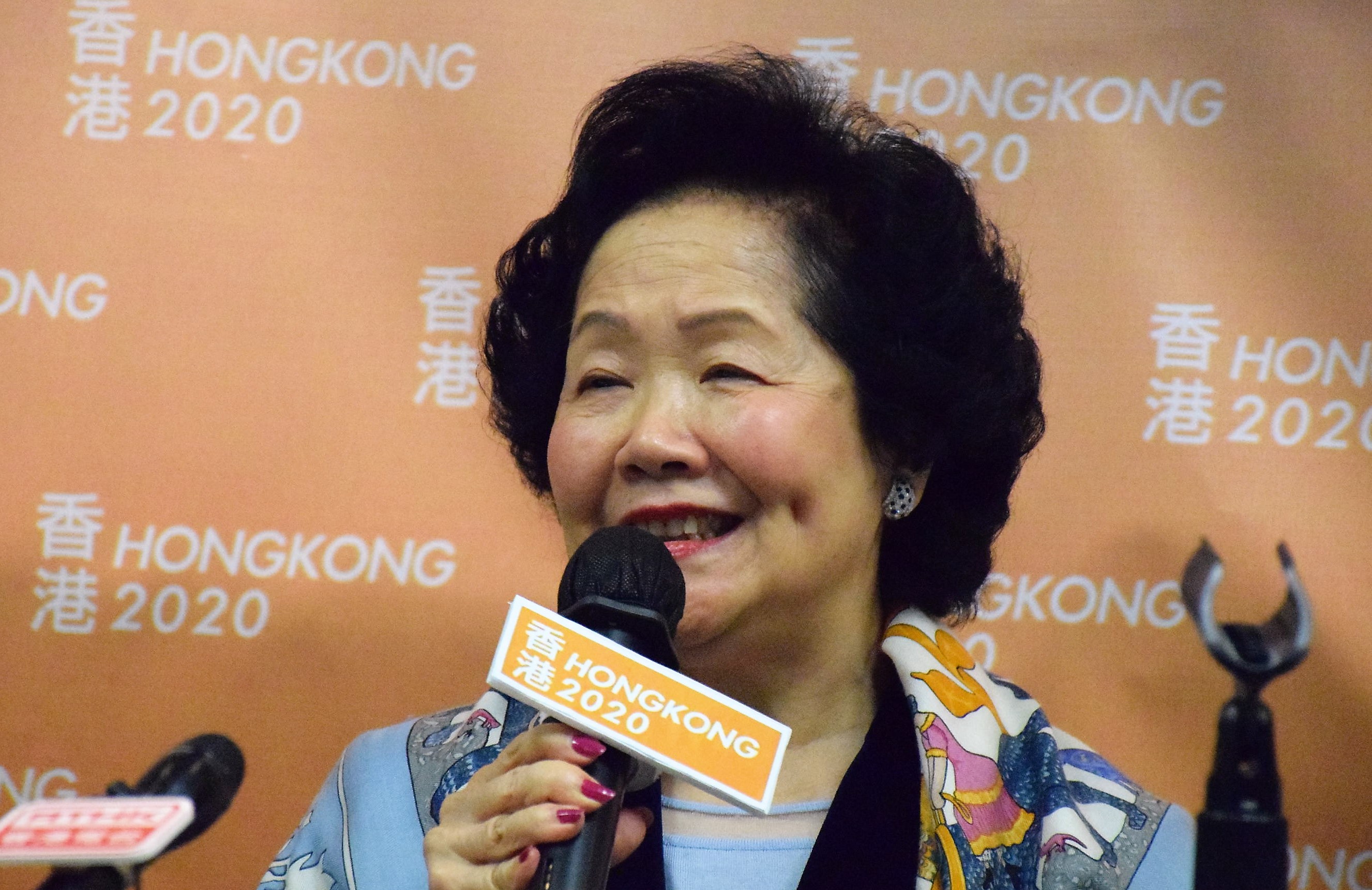 Hongkongs Chief Executive Carrie Lam bei einer Pressekonferenz, mit einem Mikrofon in der Hand und mehreren Mikrofonen vor ihr sowie einem orangen Banner im Hintergrund.