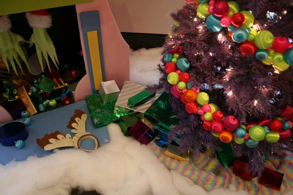 Eine Indoor-Szene mit einer Wand im Hintergrund, einem Baumwoll- und Wolltuch am Boden, einem geschm├╝ckten Weihnachtsbaum rechts, Geschenkboxen und Deko-Artikeln in der Mitte und einer H├╝tte mit Deko-Artikeln und Baumwolle ├Âber links.