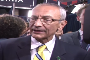 Ein Mann in Anzug und Krawatte, mit Brille, spricht zu Reportern mit einem Mikrofonständer vor ihm, umgeben von einer Gruppe von Menschen, mit Lichtern und einer Tafel mit Text im Hintergrund.